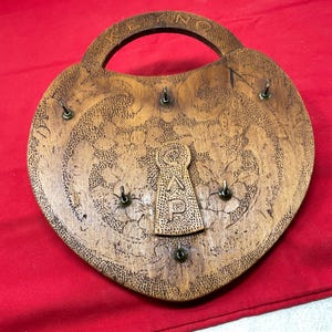 May include: Heart-shaped wooden key holder with a handle, featuring floral carvings and a keyhole design. The key holder has six metal hooks for keys and the word "CAP" is carved into the keyhole. The wood has a warm, natural finish.