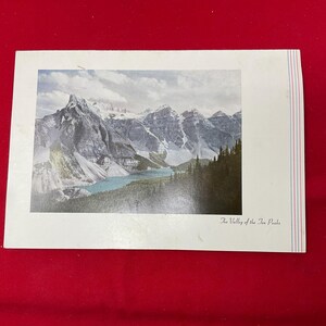May include: A rectangular postcard featuring a landscape photograph of a mountain range with snow-capped peaks and a turquoise lake. The text "The Valley of the Ten Peaks" is printed in the bottom right corner, with thin blue and red vertical lines.