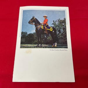 May include: A vintage paper print featuring a Royal Canadian Mounted Police officer in a red tunic and wide-brimmed hat, mounted on a black horse. The image includes a blue border and the text The Royal Canadian Mounted Police printed below.
