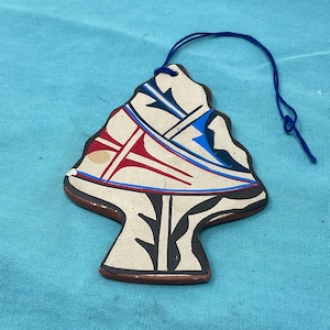 May include: A ceramic ornament in the shape of a tree with a blue string for hanging. The ornament features a cream-colored background with red, blue, and black geometric designs. The edges are brown.