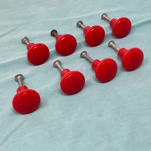 May include: A set of seven round cabinet knobs featuring a glossy red finish and a smooth, circular face. Each knob is attached to a silver-toned metal screw post, providing a simple and functional design for furniture hardware applications.