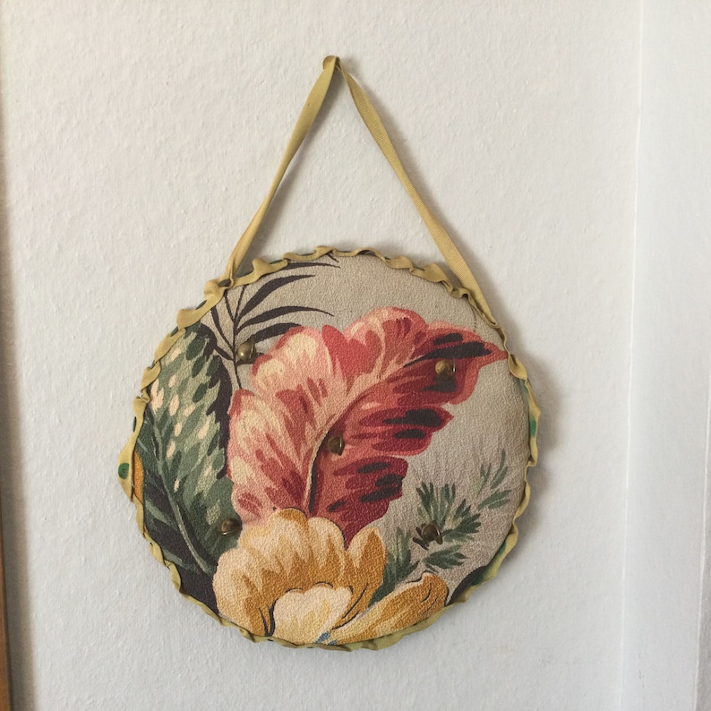 K&ouml;nnte beinhalten: Ein runder, beiger Stoff-Nadelkissen mit einem Blumendesign. Der Stoff zeigt ein gro&szlig;es rot-pinkfarbenes Blatt, eine gelbe Blume und gr&uuml;nes Laub. Das Nadelkissen ist an einer gelben Schleife zum Aufh&auml;ngen befestigt.