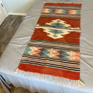 May include: A rectangular, woven runner rug with a Southwestern design. The rug features a rust-colored background with geometric patterns in teal, cream, and purple. It has fringed edges and is likely made of natural fibers.