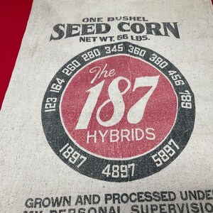 May include: A vintage seed corn sack with the text "ONE BUSHEL SEED CORN NET WT. 56 LBS." The sack features a large red circle with "The 187 HYBRIDS" and a black ring with numbers.
