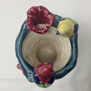 Op de afbeelding: Een keramische plantenpot in de vorm van een boomstam met drie vogels die op de takken zitten. De vogels zijn geschilderd in tinten blauw, geel en rood. De pot heeft een witte binnenkant en een blauwe en groene buitenkant.