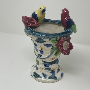 Op de afbeelding: Een witte keramische vaas met een blauw, groen en rood bloemenontwerp. Twee vogels, een gele en een rode, zitten op de rand van de vaas.