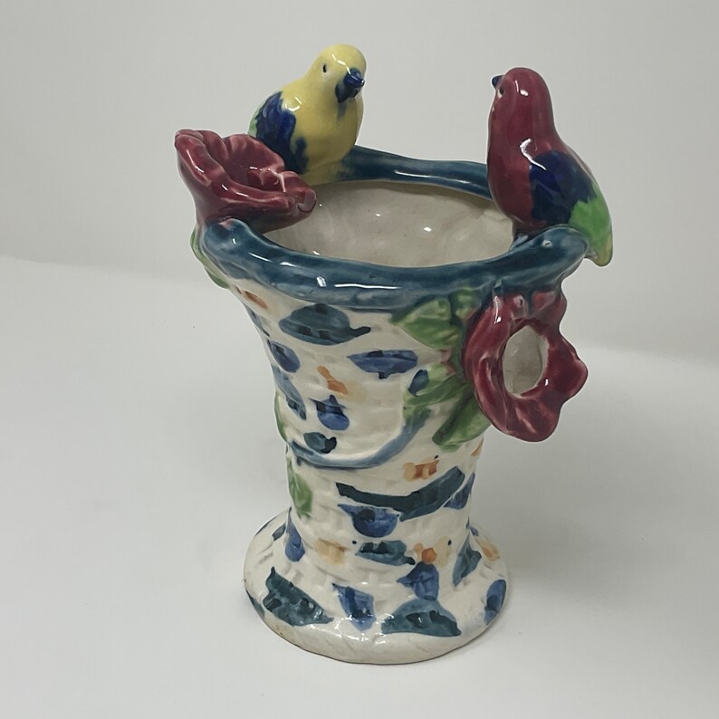 Op de afbeelding: Een witte keramische vaas met een blauw, groen en rood bloemenontwerp. Twee vogels, een gele en een rode, zitten op de rand van de vaas.