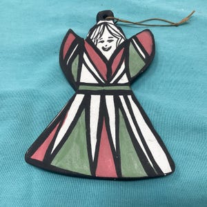 May include: Hand-painted ceramic angel ornament with a smiling face, black outlines, and a geometric design in red, green, and white. The angel has a loop for hanging and is a festive holiday decoration.