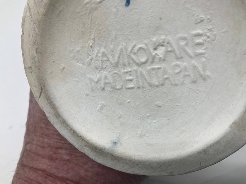 Op de afbeelding: Close-up van de onderkant van een witte keramische vaas met de tekst "Hawkoare Made in Japan" in zwart gestempeld.