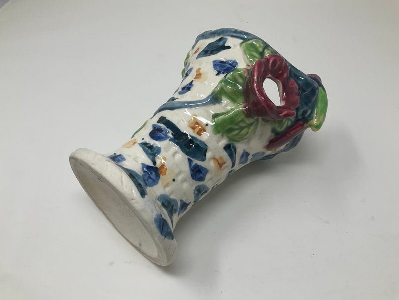 Op de afbeelding: Een witte keramische vaas met een blauw, groen en rood bloemenontwerp. De vaas heeft een brede opening en een smalle basis.