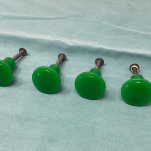 May include: Four round, bright green cabinet knobs with a smooth, glossy finish. Each knob features a central, circular top surface and a dark metal screw stem extending from the back, designed for attachment to furniture or cabinetry doors.