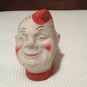 May include: A white ceramic figurine of a smiling man with red painted accents on his hair, cheeks, and collar. The figurine has a whimsical and cartoonish style.