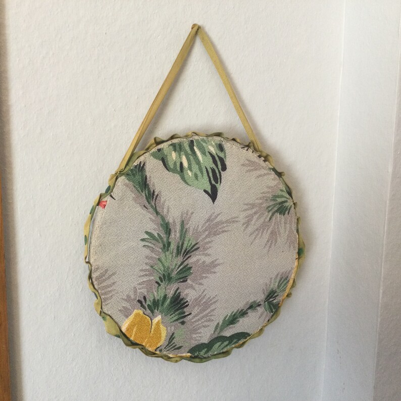 K&ouml;nnte beinhalten: Ein runder, stoffbezogener Wandbehang mit einem floralen Muster in Gr&uuml;n-, Gelb- und Graut&ouml;nen. Der Stoff ist an einer gelben Schleife zum Aufh&auml;ngen befestigt.