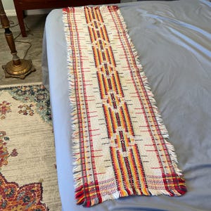 May include: A rectangular, woven table runner with a cream-colored background and a geometric pattern in red, yellow, and orange. The runner has fringed edges and is displayed on a light blue surface.