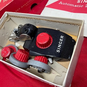 May include: A black Singer sewing machine attachment with a prominent red dial and white lettering, accompanied by three additional red and grey circular components, all housed within a rectangular cardboard box with internal dividers.