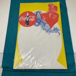 May include: Vintage Coca-Cola advertisement featuring a football player in a red uniform. The ad has a yellow background with a white oval and the Coca-Cola logo. The image is encased in plastic.