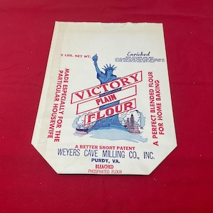 May include: Vintage Victory Plain Flour bag with a blue illustration of the Statue of Liberty. The bag is cream-colored with red and blue text, including "5 LBS. NET WT." and "A PERFECT BLENDED FLOUR FOR HOME BAKING."