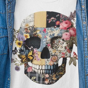 Peut inclure: T-shirt blanc avec un motif de crâne floral. Le crâne est composé de divers motifs floraux, dont des roses, des papillons et du tartan. Une veste en jean bleue est suspendue au-dessus du t-shirt, créant un look décontracté et superposé. Le t-shirt est présenté sur un cintre sur fond gris.