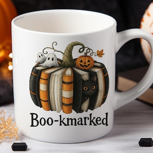 K&ouml;nnte beinhalten: Wei&szlig;e Keramiktasse mit einem Halloween-Design. Das Design zeigt einen K&uuml;rbis aus B&uuml;chern, einen K&uuml;rbis, Geister und eine schwarze Katze. Der Text "Boo-kmarked" ist auf der Vorderseite aufgedruckt.