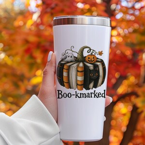 K&ouml;nnte beinhalten: Wei&szlig;er Edelstahlbecher mit einem Halloween-Motiv. Das Design zeigt einen K&uuml;rbis aus B&uuml;chern, eine schwarze Katze, Geister und eine Jack-o'-Laterne. Der Text "Boo-kmarked" ist darunter gedruckt.
