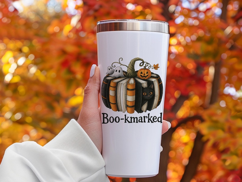 K&ouml;nnte beinhalten: Wei&szlig;er Edelstahlbecher mit einem Halloween-Motiv. Das Design zeigt einen K&uuml;rbis aus B&uuml;chern, eine schwarze Katze, Geister und eine Jack-o'-Laterne. Der Text "Boo-kmarked" ist darunter gedruckt.