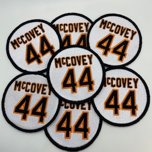 May include: Seven white, round, embroidered patches with black outlines. Each patch features the name "McCovey" and the number "44" in orange with a black outline.