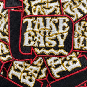 May include: Black embroidered patches with the phrase "TAKE IT EASY" in white and gold thread. The patches have a red border and are scattered across the image. The patches are likely for clothing or accessories.