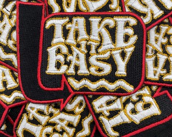 May include: Black embroidered patches with the phrase "TAKE IT EASY" in white and gold thread. The patches have a red border and are scattered across the image. The patches are likely for clothing or accessories.
