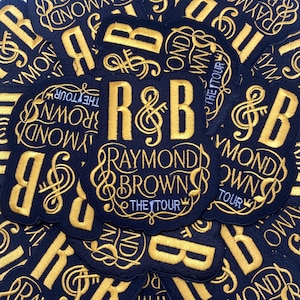 May include: A collection of navy blue embroidered patches. Each patch features the letters "R&B" with a treble clef, the name "Raymond Brown", and the words "The Tour" in gold thread.
