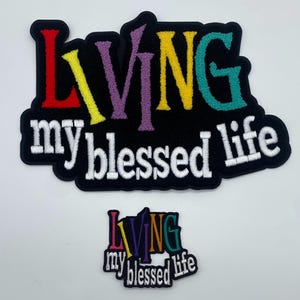 May include: Two embroidered patches with the text "Living my blessed life" in white lettering on a black background. The larger patch has the word "Living" in rainbow colors. The smaller patch has the word "Living" in rainbow colors and is slightly smaller.
