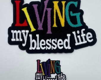 Könnte beinhalten: Zwei gestickte Aufnäher mit dem Text "Living my blessed life" in weißen Buchstaben auf schwarzem Hintergrund. Der größere Aufnäher hat das Wort "Living" in Regenbogenfarben. Der kleinere Aufnäher hat das Wort "Living" in Regenbogenfarben und ist etwas kleiner.
