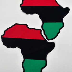 May include: Two fabric patches in the shape of the African continent. Each patch features a red stripe at the top, a black stripe in the middle, and a green stripe at the bottom, with a black border. The patches are placed on a white background.