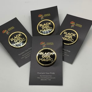 Op de afbeelding: Vier zwarte en gouden emaille-pins met de tekst "Black Girl Magic" op een zwarte achtergrond. De pins bevinden zich op zwarte kaarten met de tekst "Proclaim Your Pride" en de bedrijfsnaam "Prideful Patchez".