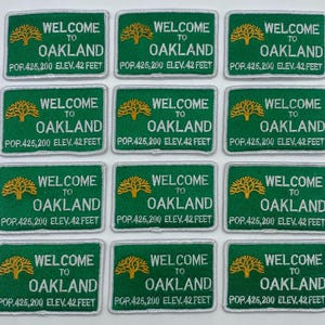 May include: A collection of green embroidered patches. Each patch features the words "WELCOME TO OAKLAND" in white, with a gold tree graphic. The text "POP.425,200 ELEV. 42 FEET" is also included.