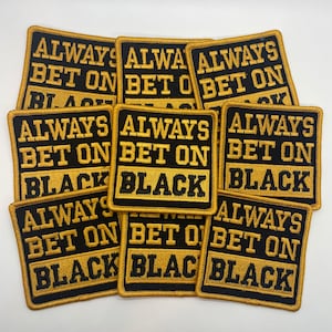 Op de afbeelding: Een stapel van negen zwarte en gouden geborduurde patches met de tekst "Always Bet On Black".