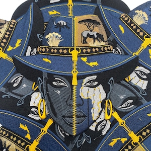 以下が含まれることがあります： 女性の顔とアフリカのサバンナのイメージが刺繍された青と黒の帽子。帽子は中央にジッパーのデザインと金色のアクセントが特徴です。デザインには、象、木、幾何学模様が含まれています。