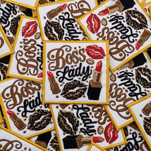 Puede incluir: Parches bordados blancos con borde dorado, con el texto "Boss's Lady" en escritura marrón. Los parches también incluyen lápiz labial, labios con estampado de leopardo y diseños de granos de café. Un accesorio con temática de belleza.