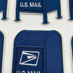 May include: A collection of embroidered patches shaped like blue U.S. Mail collection boxes. Each patch features a dark blue background with white embroidered details, including the U.S. Mail logo and the text U.S. MAIL, finished with a cream border.