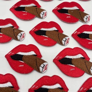 May include: Embroidered patches featuring red lips with a brown cigar. The lips are open, revealing a black interior and white teeth. The cigars have a detailed design, with a white end and a brown body. The patches are arranged on a white surface.