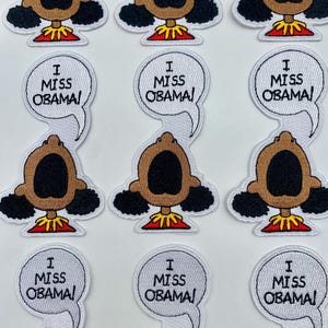 May include: Nine white embroidered patches with a cartoon character with brown skin and black hair. The character has a large open mouth and is wearing a red and yellow outfit. Each patch has a white speech bubble with the text "I Miss Obama!"