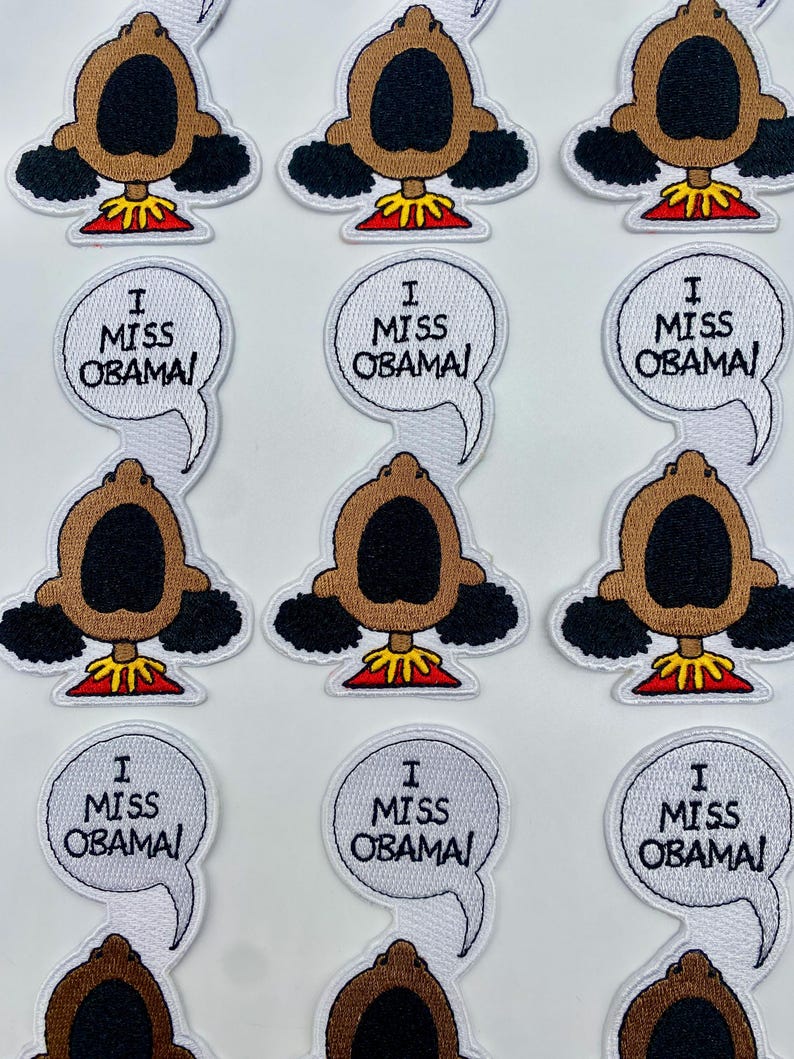 May include: Nine white embroidered patches with a cartoon character with brown skin and black hair. The character has a large open mouth and is wearing a red and yellow outfit. Each patch has a white speech bubble with the text "I Miss Obama!"
