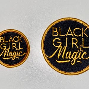 May include: Two embroidered patches with the text "BLACK GIRL MAGIC". The patches are circular, with a black background and gold lettering and border. One patch is smaller than the other.