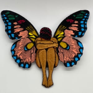 May include: A colorful embroidered patch of a person sitting with butterfly wings. The wings are pink, yellow, blue, green, and black. The person is brown and has their arms crossed.