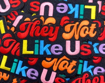 May include: A collection of embroidered patches with the phrase "They Not Like Us" in a variety of colorful fonts. The patches have a black background and are in various sizes and orientations.