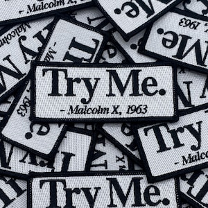 May include: A collection of rectangular white fabric patches with black embroidered borders and text. Each patch features the words Try Me. followed by - Malcolm X, 1963 in a bold serif font. The patches are layered in a dense, overlapping pile.
