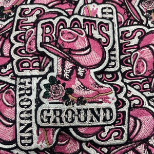 以下が含まれることがあります： カウボーイブーツと帽子のデザインが施された、ピンクと白の刺繍パッチ。パッチには、装飾的なフォントで「Boots on the Ground」という言葉と花のアクセントが施されています。パッチは、衣類やアクセサリーに適しています。