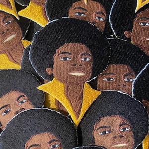 May include: A collection of embroidered patches featuring a portrait of a person with an afro hairstyle wearing a yellow shirt. The patches are made of fabric and have a black and white colour scheme.