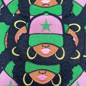 May include: A collection of embroidered patches featuring a stylized Black woman with black hair, gold hoop earrings, and a pink cap with a green star. The patches have green, pink, black, and brown colors.
