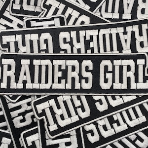 Könnte beinhalten: Nahaufnahme von mehreren schwarzen und weißen gestickten Aufnähern mit dem Text "RAIDERS GIRL" und "RAIDERS" darauf.
