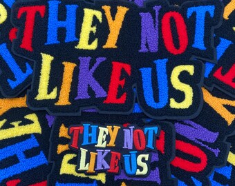 May include: A collection of black embroidered patches with the phrase "THEY NOT LIKE US" in colorful, textured lettering. The letters are in various colors, including red, blue, yellow, orange, and purple.